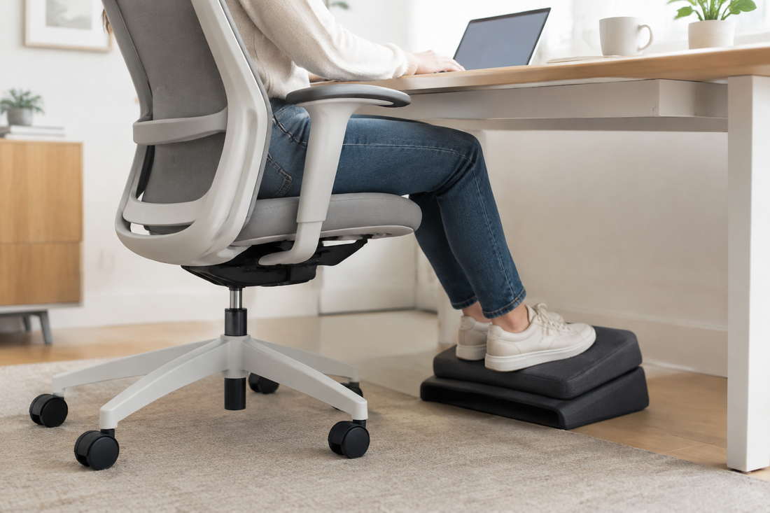 A seated office worker rests both feet on a foam under-desk footrest beneath a high office chair.